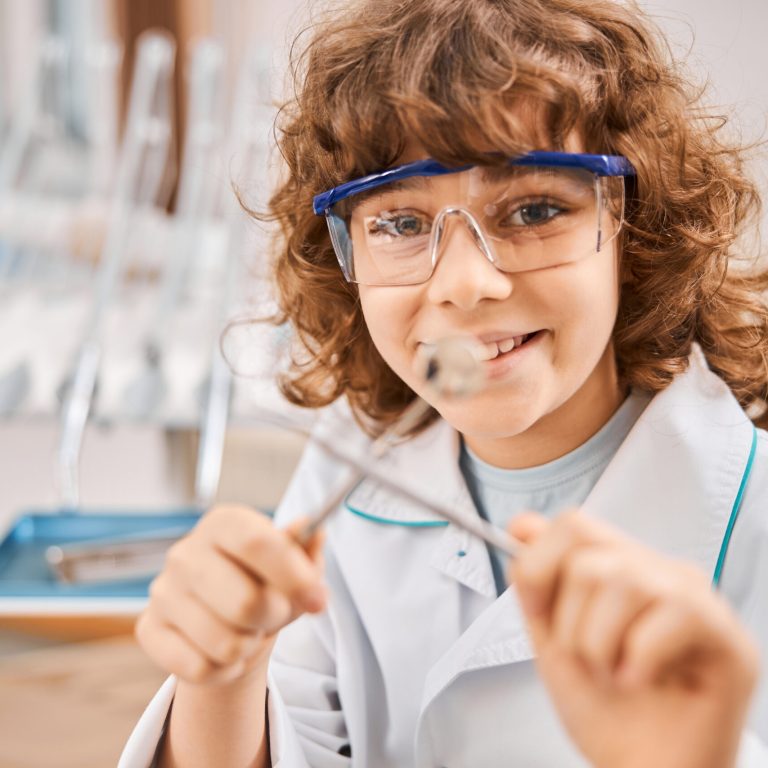 Close-up photo of a joyful kid wearing a lab coat and holding dental tools while posing in dentist office