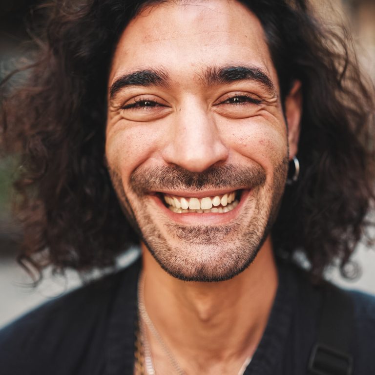 Young attractive italian guy with long curly hair and stubble looks into the camera and smiles at old buildings background. Stylish man with an earring in his ear and lot of chains