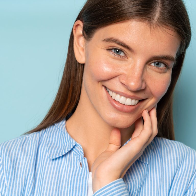 Closeup portrait of young woman with white toothy smile in casual striped shirt posing against blue studio background matching with color of her eyes touching face gently